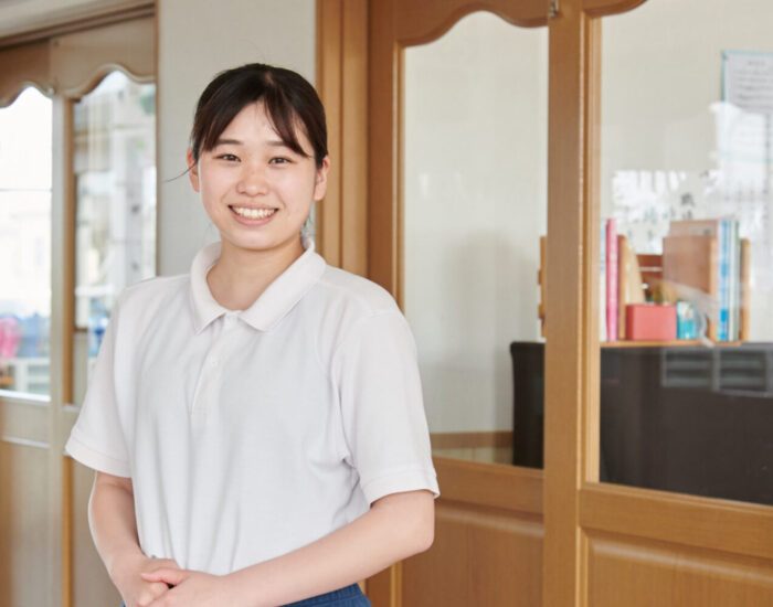 ちくみ幼稚園の園舎内、本棚が見えるガラス戸の前でほほえむみさき先生