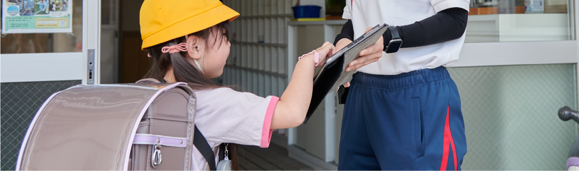 ランドセルを背負った女の子が、アフタースクールの受付するようす